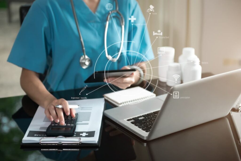 Nurse on a laptop with a tablet in her hand. In her other hand, she's using a calculator that's on a clipboard